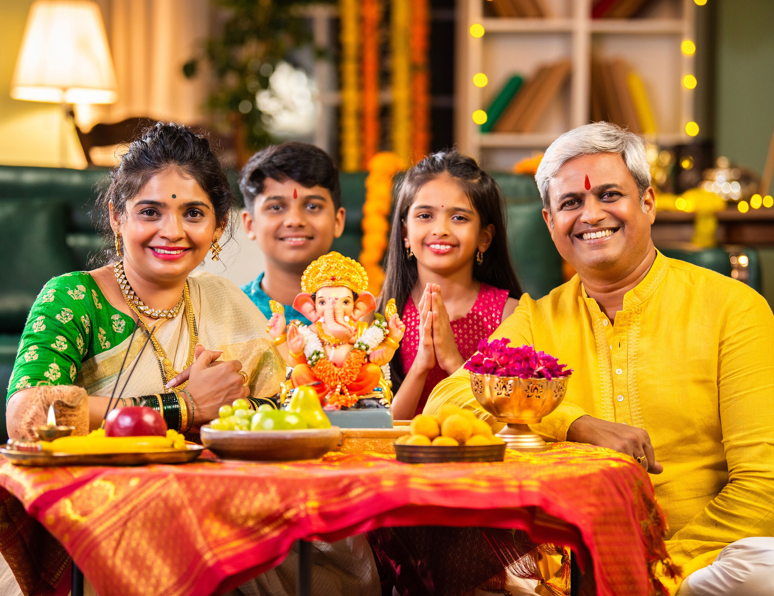 Smiling Indian family celebrating Ganesh Chaturthi at home with Lord Ganesha idol, fruits, sweets, and festive decorations